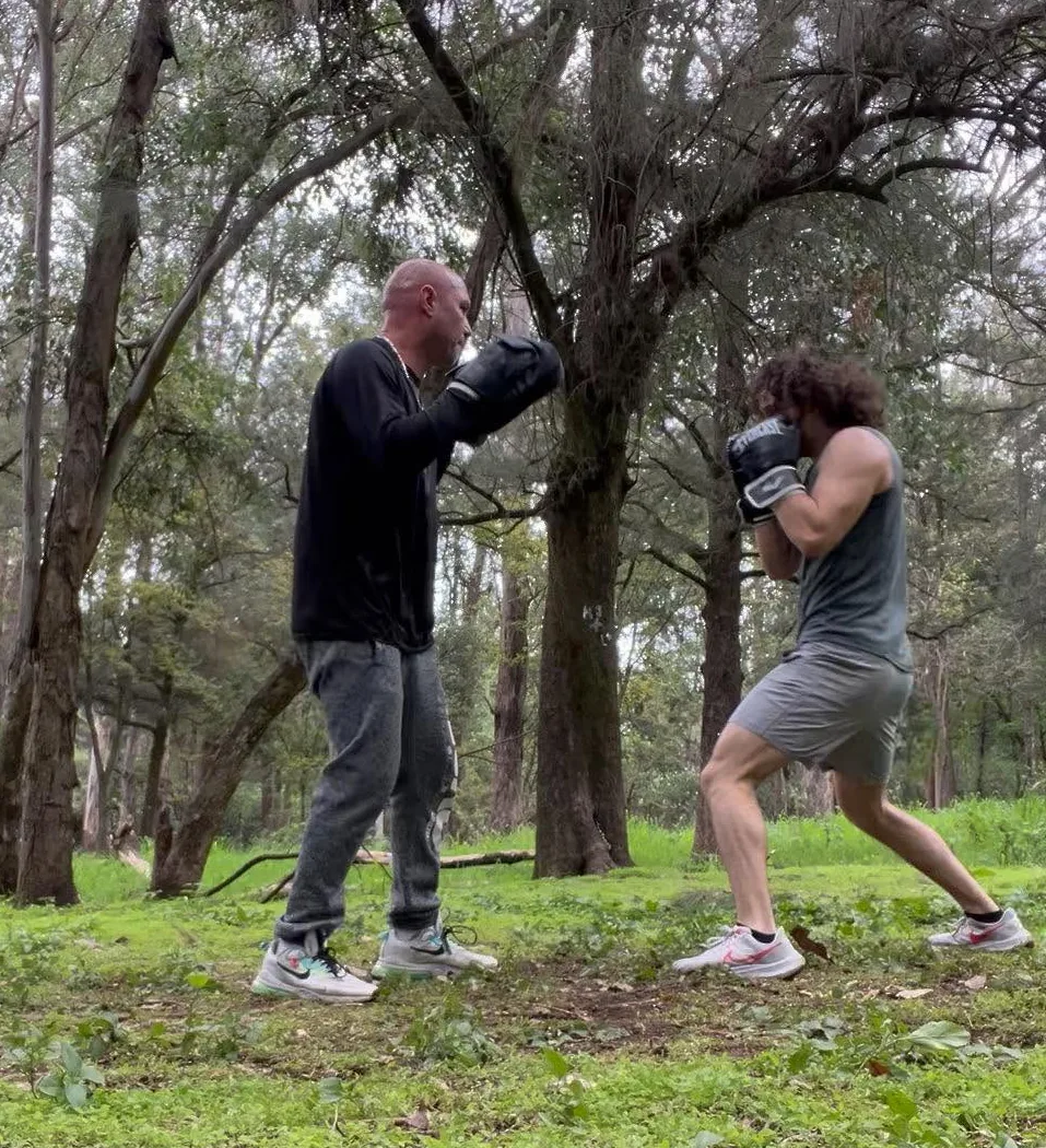 Coach and client boxing outdoors on grass among trees during a pad-work session in a Guadalajara park.